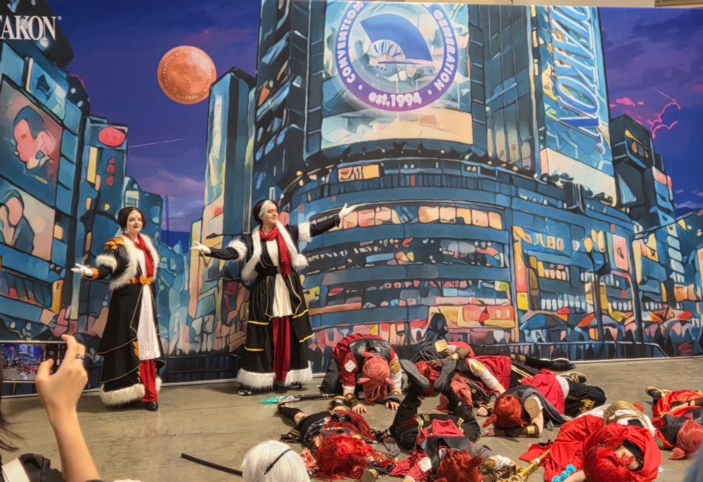 Two Emet Selch cosplayers standing triumphantly over a large gaggle of Graha Tia cosplayers laying on the floor for REASONS dw about it. 