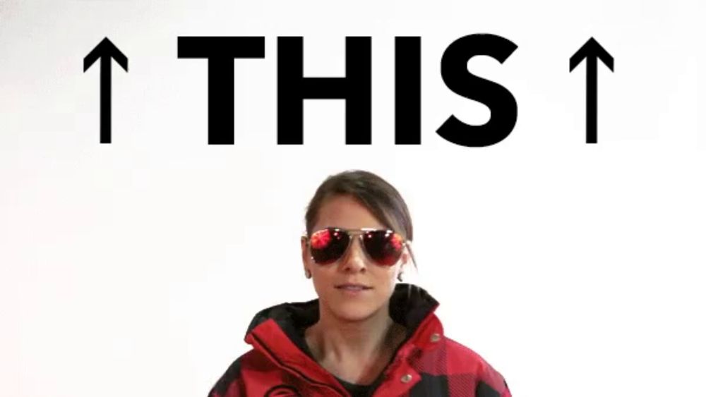 a woman wearing sunglasses and a red jacket is pointing up with the words " this " above her