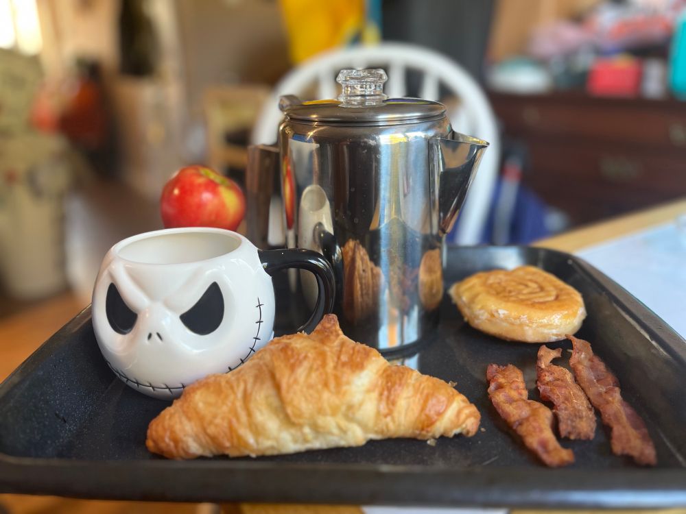 A breakfast on a tray consisting of a pot of coffee, an apple, a croissant, a sweet roll, and three strips of bacon.