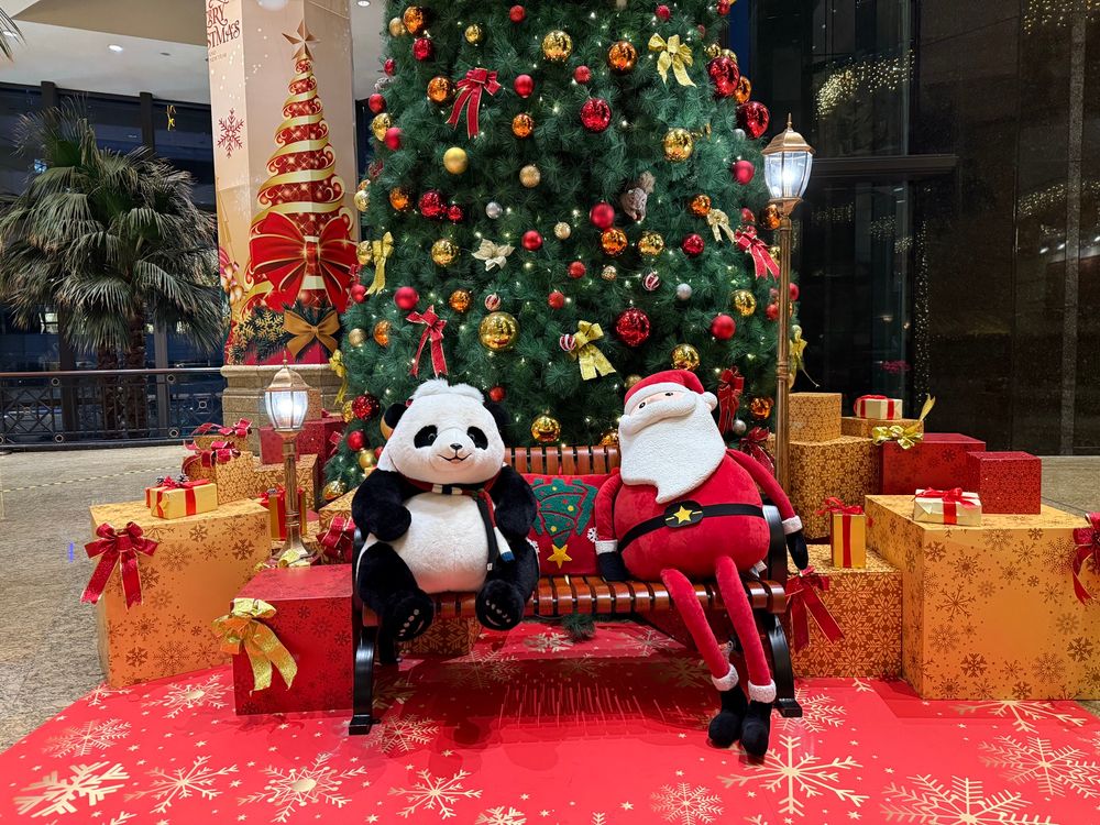 A Santa and a panda are sitting on a bench and relaxing in front of a giant Christmas tree decorated with red and gold ornaments.