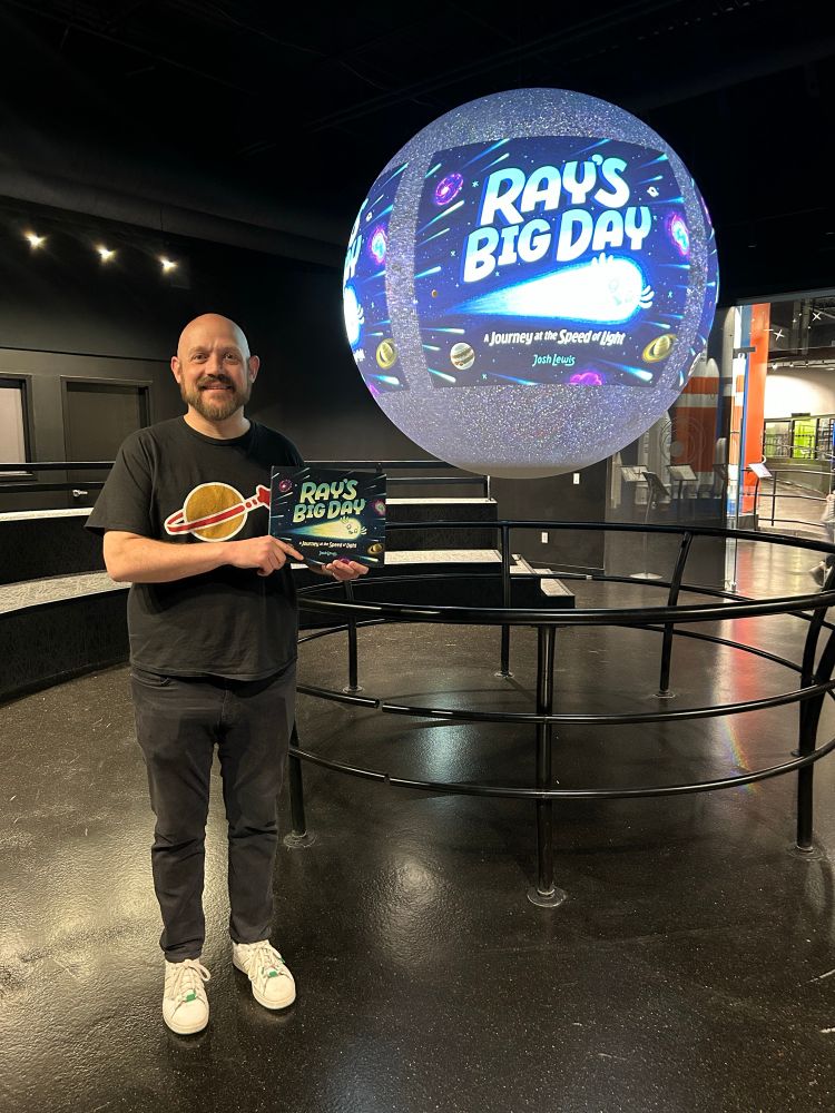 Josh Lewis holding his picture book, Ray’s Big Day, while standing in front of the Science on a Sphere at the Space Foundation Discovery Center.
