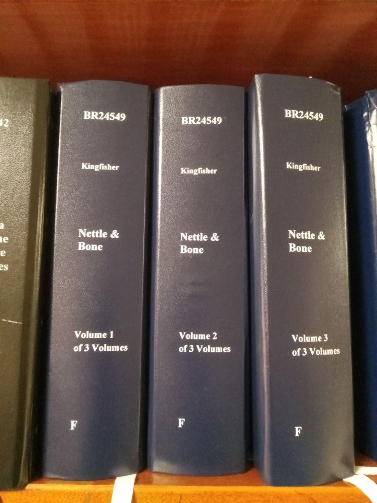 Three thick upright volumes bound in dark cloth, labeled with call number, volume number, the letter F, the name Kingfisher, and the title "Nettle and Bone."