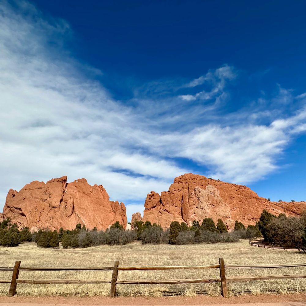 A vibrant landscape featuring striking red rock formations rising dramatically against a bright blue sky. The foreground showcases a golden grassy field bordered by a rustic wooden fence, with scattered green trees adding contrast. Wispy white clouds stretch across the sky, enhancing the natural beauty and depth of the scene. The photo captures the grandeur and ruggedness of the red sandstone formations, a hallmark of a desert or arid environment.