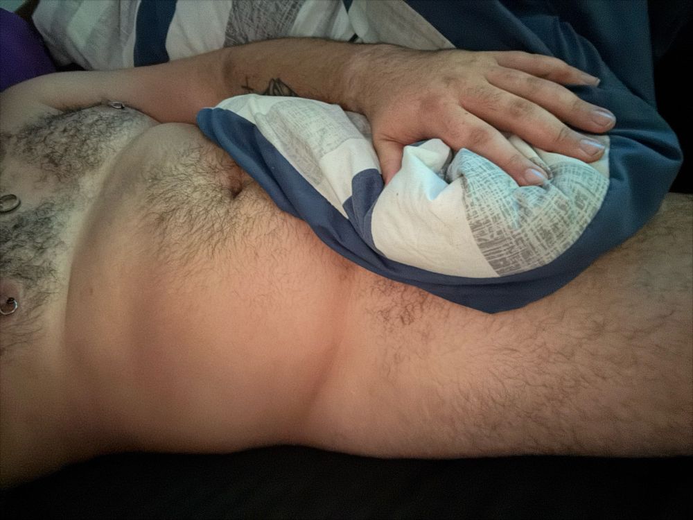 Ghost lying in bed naked with the blue & white comforter covering his lap with his chest/stomach and most of his thigh exposed