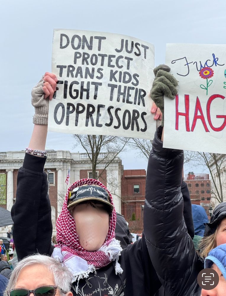 Young person hollering, holding up a sign that says “don’t just protect trans kids - Fight their Oppressors!” A woman beside them holds a sign with hand-drawn flowers that says, in cursive, “Fuck the Patriarchy - HAGS UNITE.”