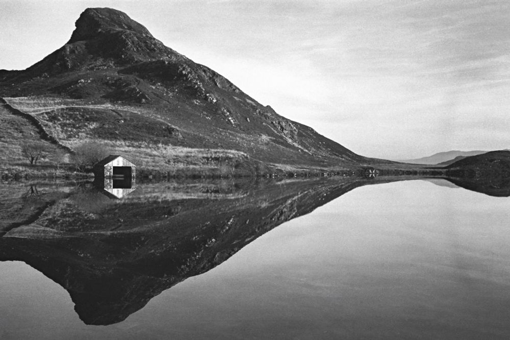 A black and white medium format film photo of a peaked Welsh hill that’s not quite a mountain, and it’s almost identical reflection in a still lake below it. There’s also a small boat house on the lake shore. 