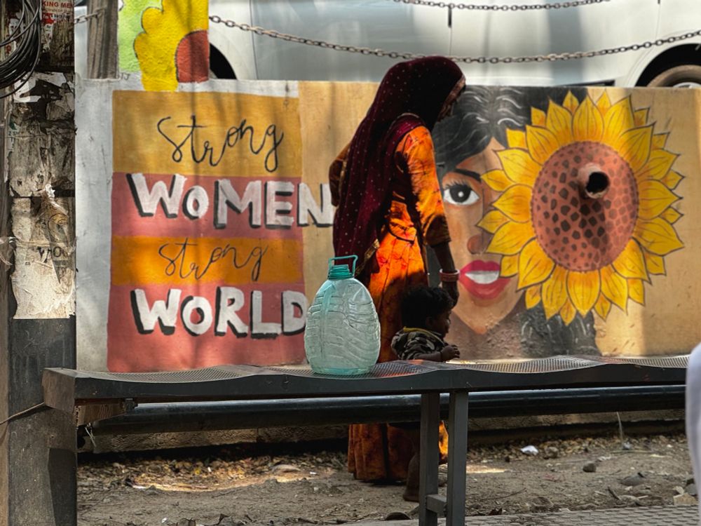 A woman holding her child’s hand on the side of a street. They are standing in front of a wall with a mural of a woman’s face next to a sunflower, with the writing “strong women, strong world”
