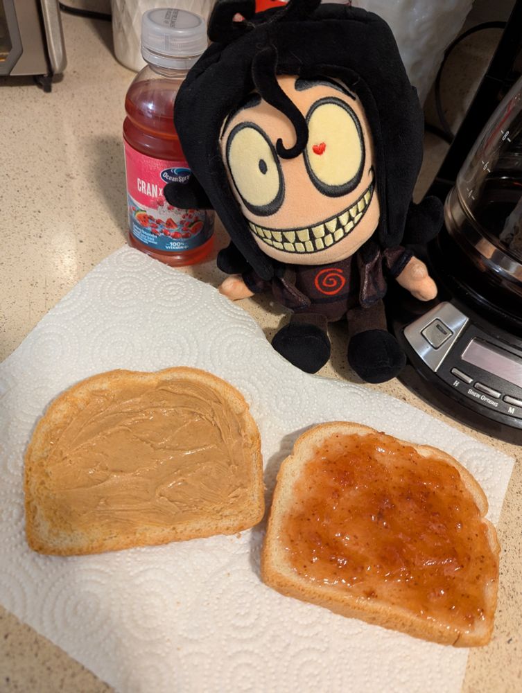 Slices of bread with peanut butter n strawberry jelly behind a plushie of John Doe