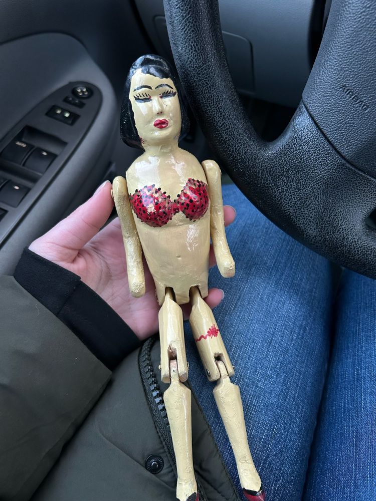 A wood-carved doll with jointed legs of a white pin-up woman with black hair, red lips, a red bra and garter, and red heeled shoes. 