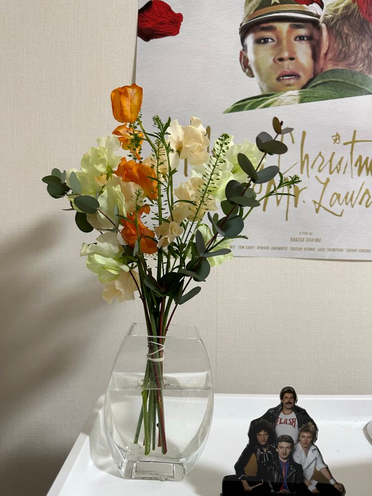 白いラックの上に花瓶にいけられた花束、クイーンのアクリルスタンドが置いてあり、背景の壁には映画『戦場のメリークリスマス』のポスターが貼ってある