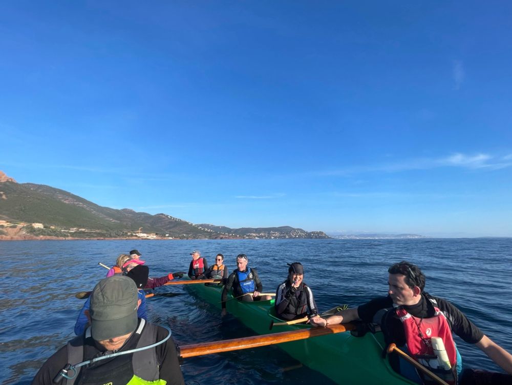 Paddlers on tauati va’a in the foreground, the red volcanic rocks of the Esterel massif, and the snowy summits of the Mercantour massif in the back