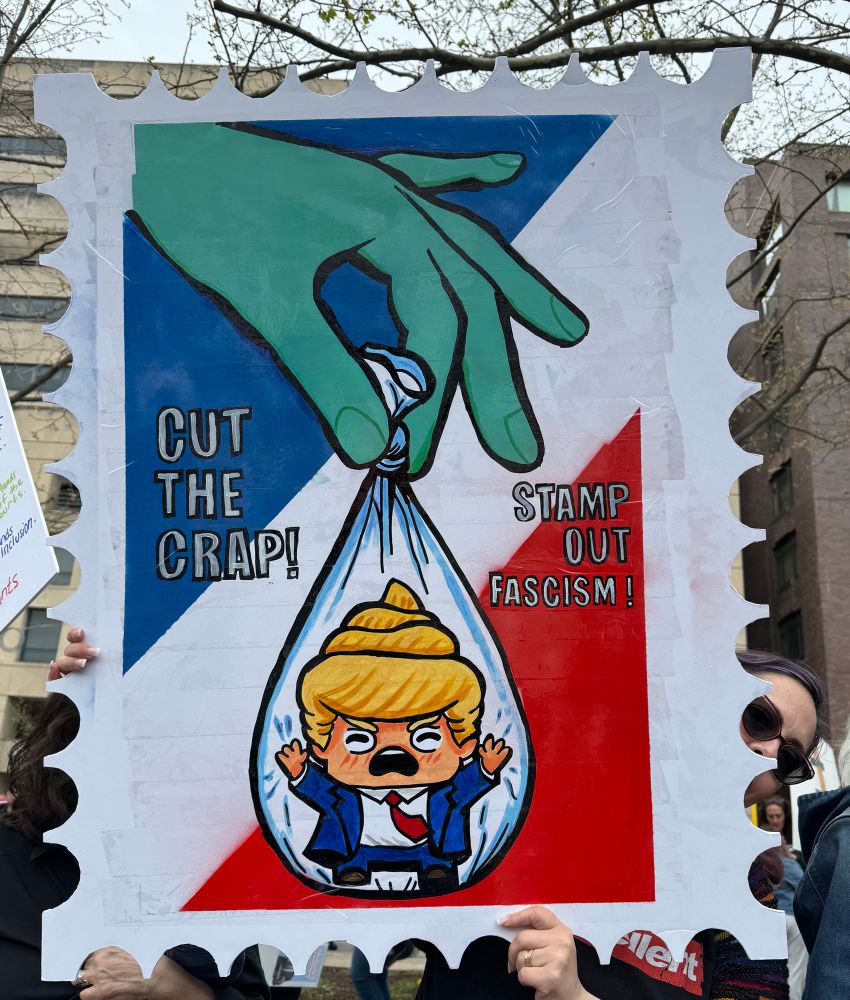  Graphic sign in the shape of a postage stamp showing a crying baby resembling Don Trump, in a clear bag. The bag is held by a hand with green fingers. The sign says “Cut the crap” and “Stamp out Fascism” 