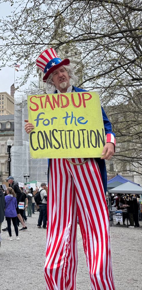 Picture of a man on stilts, dressed as Uncle Sam, holding a sign that says “Stand up for the Constitution”