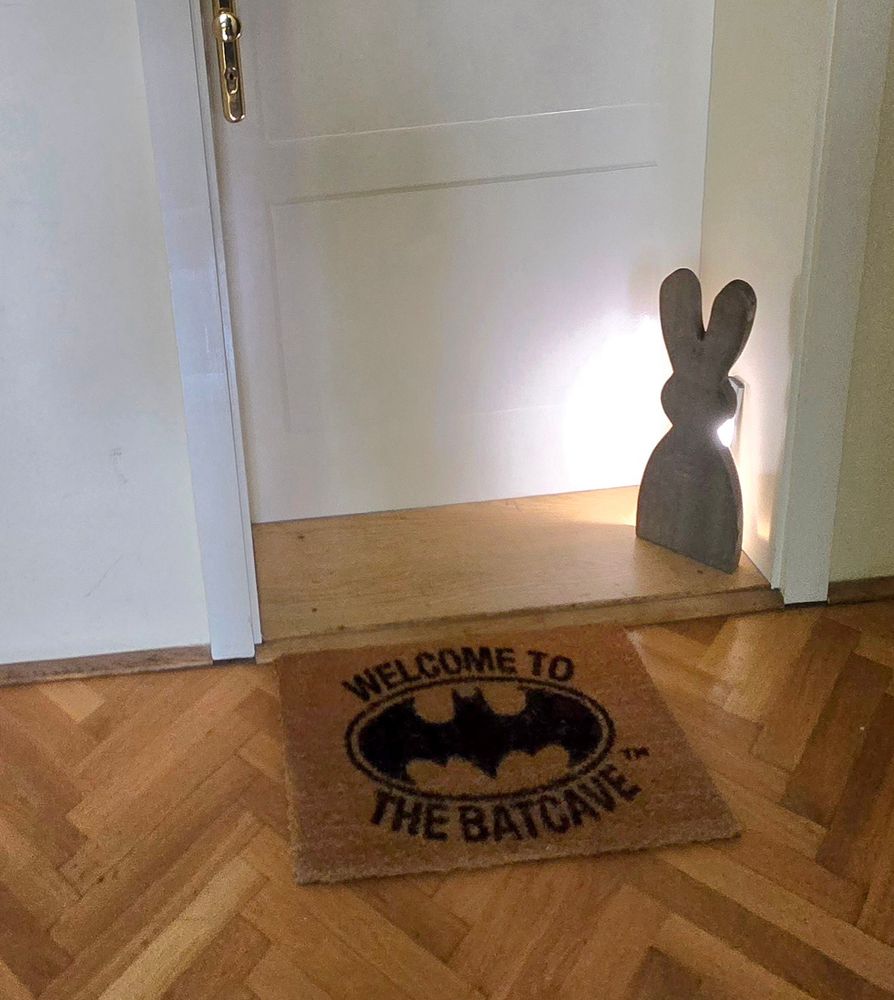 Das Bild zeigt einen Wohnungseingang, eine weiße Tür. Am etwas 30cm tiefen Türantritt, steht eine ca. 40cm große grau-braune Osterhasenfigur aus Holz, die von hinten angeschienen wird.
Davor sieht man den Stab-Parkett des Hausflures, der im Fischgrät-Muster verlegt wurde. Auf dem eine Fussmatte liegt, das Batman-Logo zeigt, außerhalb läßt sich der Spruch, 'Welcome to the Batcave' ablesen.