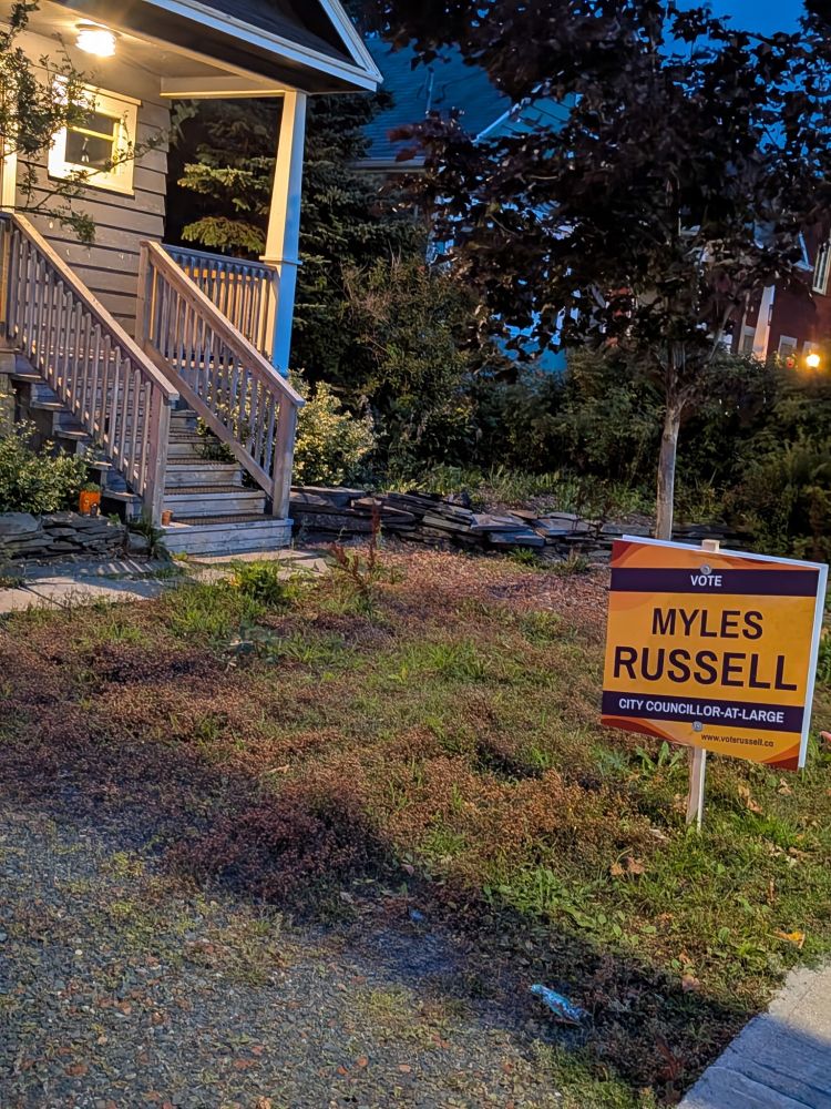 Vote Myles Russell City Councillor At-Large sign; purple lettering on golden yellow background. Red Maple tree behind Sign next to dark grey house with wood steps and veranda.