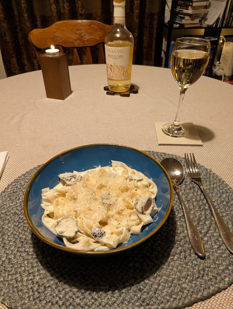 Dining table with a candle, bottle of white wine Vesevo Greco Di Tufo and a glass of the same. On a woven blue tablemats there's a fork and spoon to the right. There's a blue ceramic pasta bowl with creamy black truffle tagliatelle. 