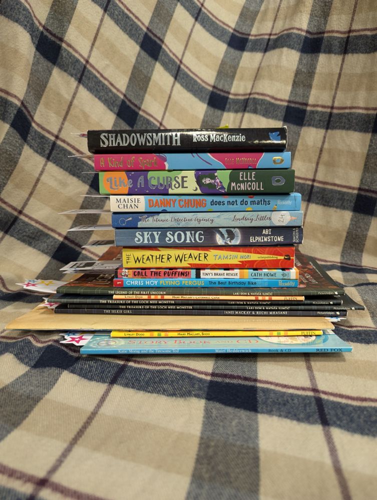  Stack of books on a plaid scarf. 
Top to bottom: 
Shadowsmith by Ross Mackenzie 
A Kind of Spark by Elle McNicoll
Like a Curse by Elle McNicoll
Danny Chung does not do maths by Maisie Chan
The Titanic Detective Agency by Lindsay Littleson 
Sky Song by Abi Elphinstone 
The Weather Weaver by Tamsin Mori
Call the Puffins: Tiny's Brave Rescue by Cath Howe
Flying Fergus The best birthday cake by Chris Hoy
The legend of the first unicorn by Lari Don and Natasa Ilincin 
The treasure of the Loch Ness monster by Lari Don and Natasa Ilincin 
The Selkie Girl by Janis Mackay and Ruchi Mhasane 
Hairy McClary's Caterwaul Caper by Lynley Dodd
Hairy McClary, Shoo by Lynley Dodd
Katie Morag and the tiresome Ted by Mairi Hedderwick 
The Smoozles of Orkneyjar by Sallianne Smales
 