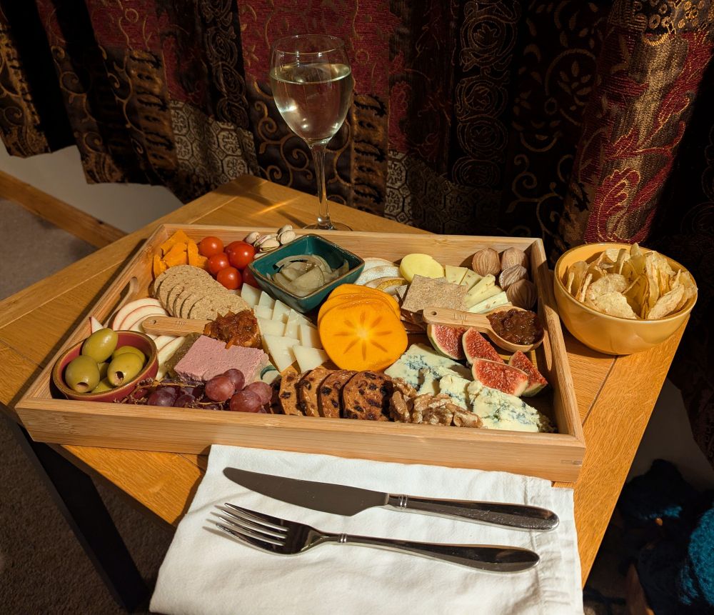 Small wooden side table with a bamboo tray on it and a white cloth napkin with knife and fork on it. There's a glass of white wine behind the tray and a small yellow ceramic bowl with crisps in to the right of it. On the tray is cashel blue cheese, Gouda with cumin, ossau iraty and Rutland red. There's a portion of pate, small square sourdough crackers, small round oatcakes (local), and Millers plum and date toasts. Fresh fig, sliced persimmon, apple and pear, red grapes, cherry tomatoes, walnuts, pistachios, pickled onions, green olives in a small round dish, pickled cucumber in a small square dish. Lastly two wooden measuring spoons with different chutneys in them. 