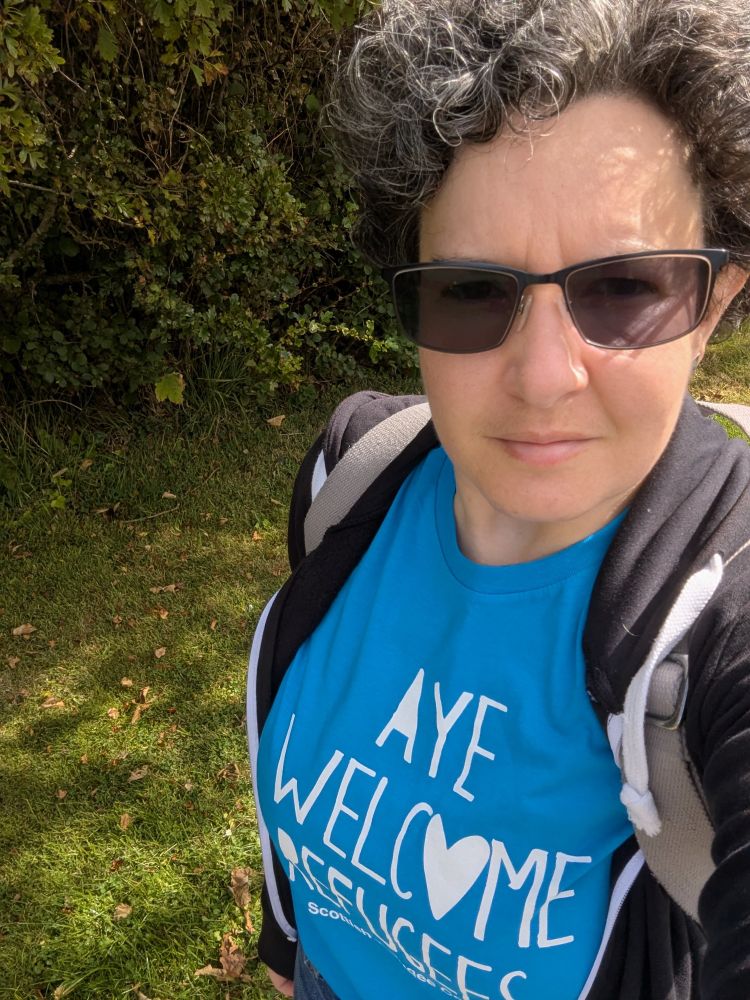 Me: short untidy curly dark brown with increasing silver stands! Glasses, black zip up hoody undone wearing my Aye Welcome Refugees t-shirt (bright blue) from the Scottish Refugee Council. 