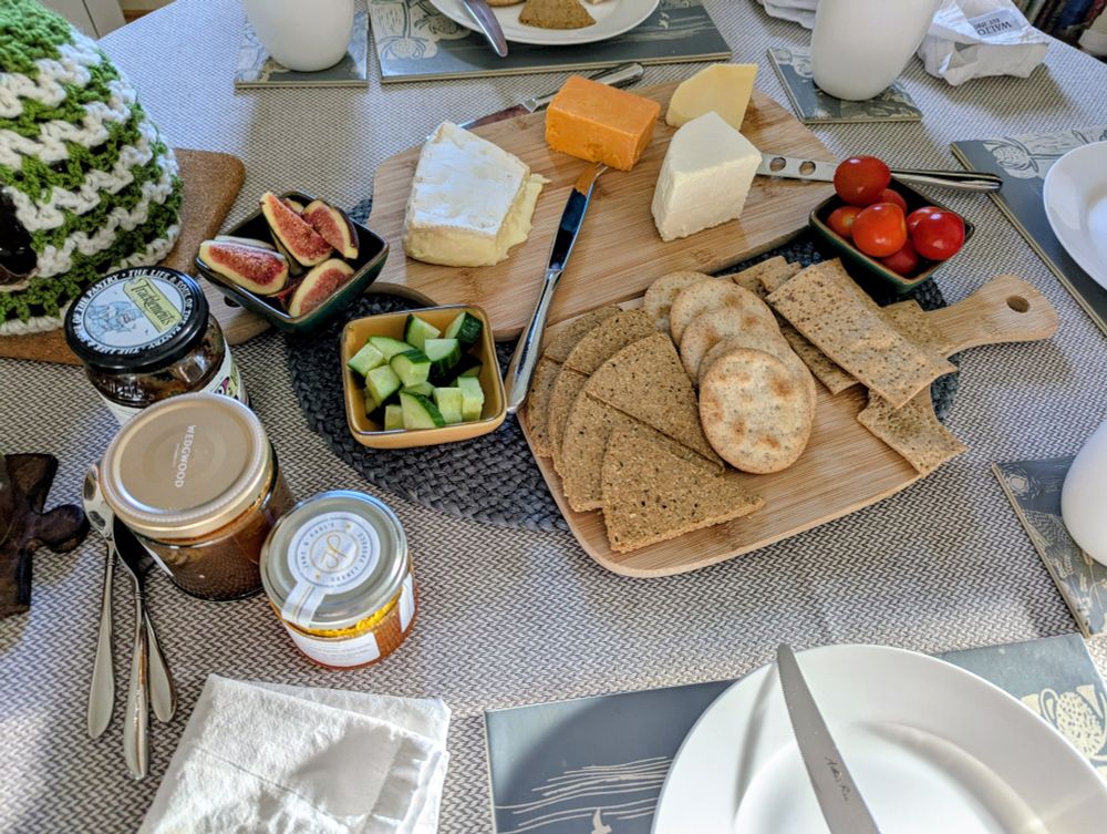 Spread of cheese and crackers on my dining table with dishes of cherry tomatoes, cucumber and fresh figs. There are 3 jars of chutney/relishes and the edge of the teapot, plates and mugs around the table. 