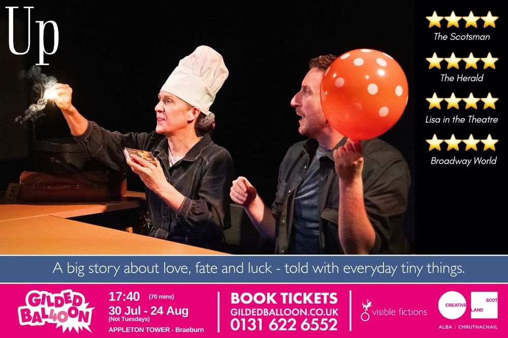 Two people sit at a desk at a dark stage. One is holding a polkadot balloon and one is wearing a chef's hat and has lit a match. 