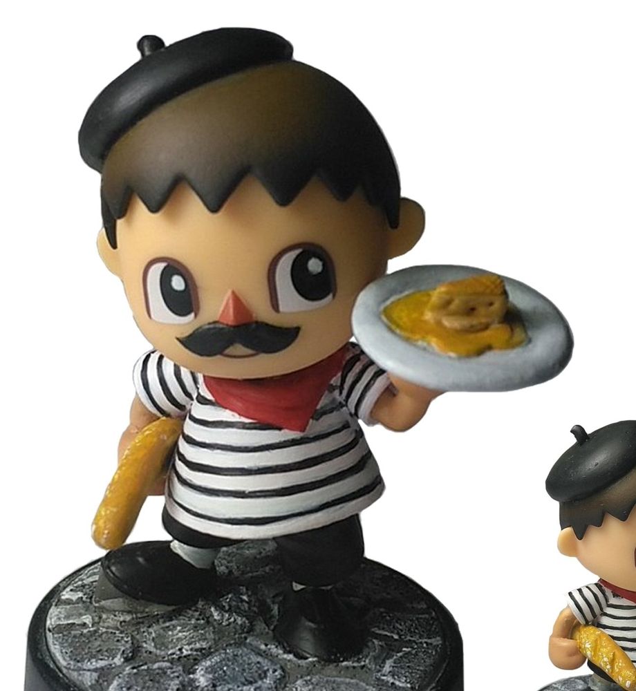 Une figurine représentant un petit bonhomme moustachu avec un béret, une marinière, un foulard rouge, une baguette sous la main et portant une assiette avec une omelette et du fromage 