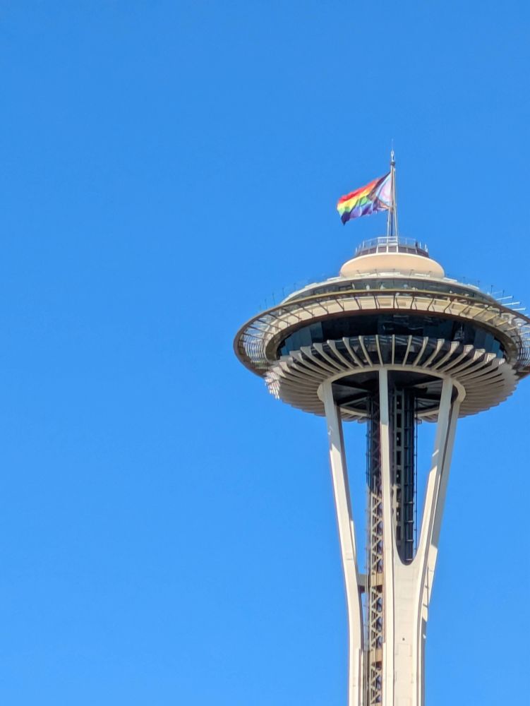 The upper portion of the Seattle Space Needle with a Progress Flag flying from the top of it. The sky behind it is a brilliant blue.