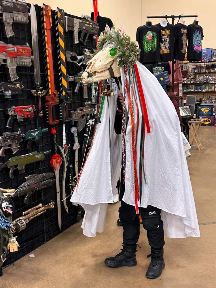 Mari Lwyd - a sort of hobby horse using a real horses skull held aloft on a pole. The human operator is covered in a white sheet, skull held aloft and decorated with colourful ribbons. Mari wears a crown of evergreen foliage. This one is standing in front of a display of replica weapons from popular sci-fi movies 