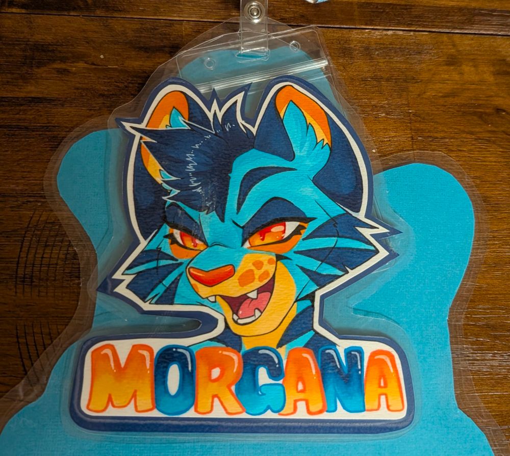 A laminated headshot badge of Morgana, a blue and orange monster cat, with a mischievous expression