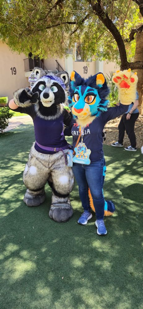 A raccoon fursuiter makes a peace sign and a blue cat fursuiter waves while standing in a hotel courtyard.