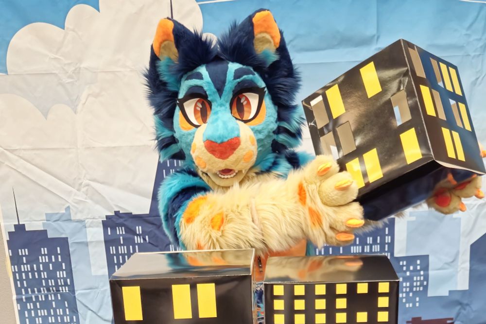 A blue cat fursuiter holds a cardboard box that resembles a piece of a skyscraper with a cityscape background 