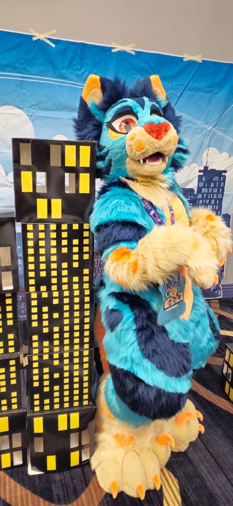 A blue cat fursuiter stands with her back against a miniature cardboard skyscraper