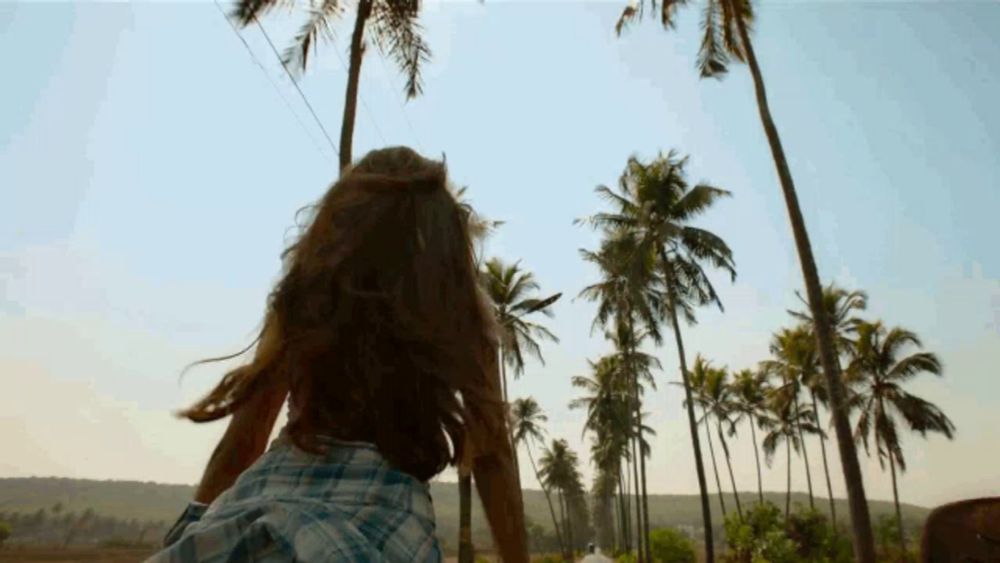 a woman in a plaid shirt is riding on the back of a man on a road with palm trees in the background