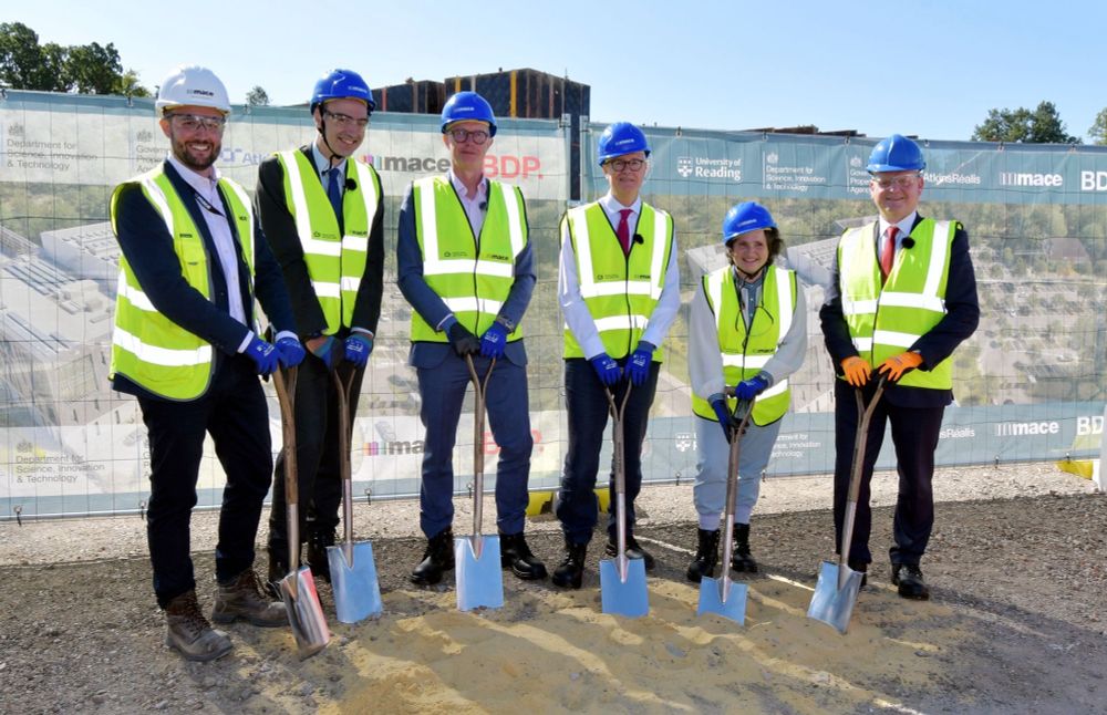 Six smiling people in hard hats and hi vis with spades behind a small hole in front of a big building with