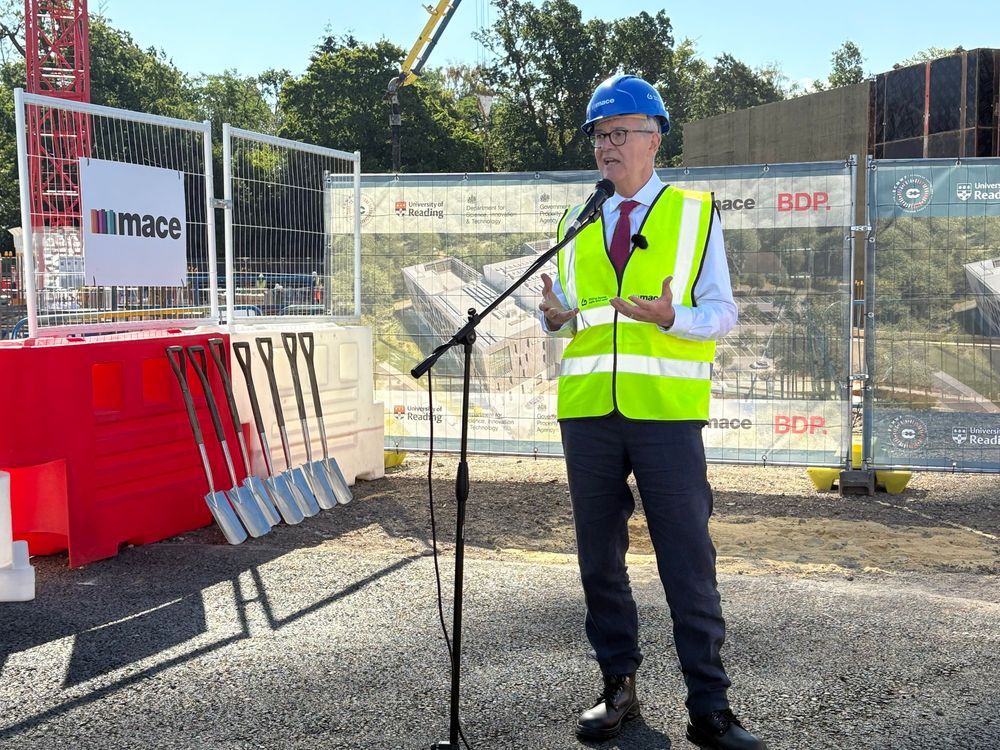Lord Vallance speaking at a microphone in full PPE with the spades lined up behind him