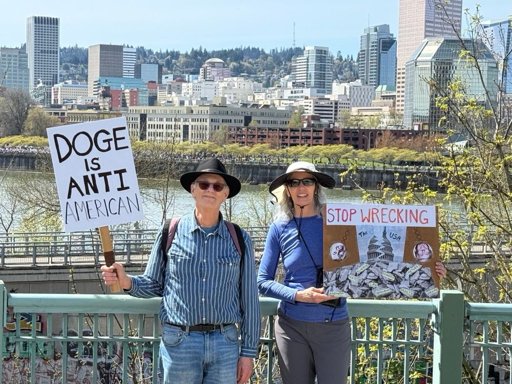 Protest signs say “DOGE is anti American “ and “stop wrecking the US” with River behind the protestors in Portland. 
