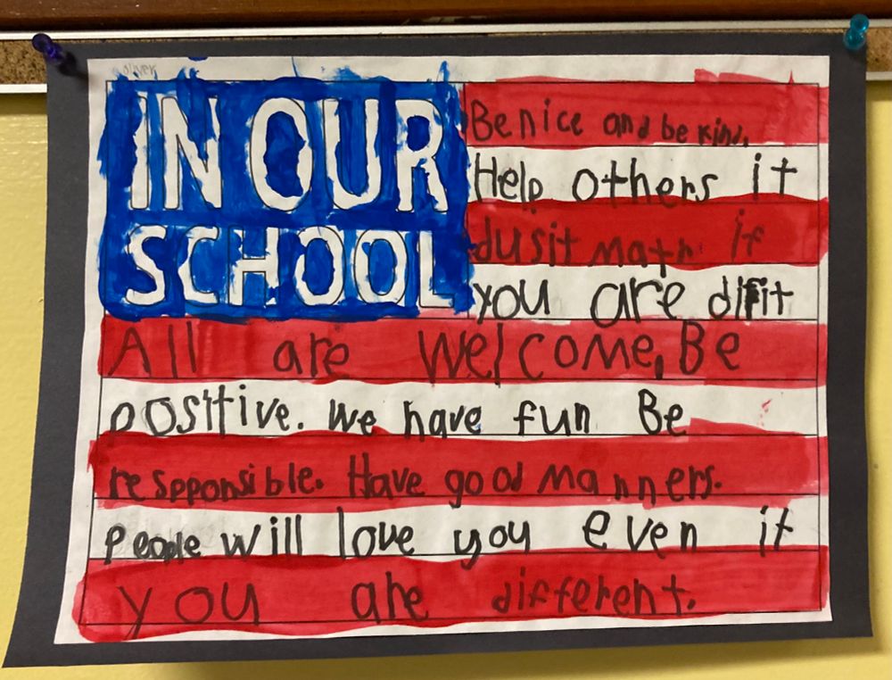 US flag with hand written words on it. In our school be nice and be kind. Help others. It doesn’t matter if you are different. All are welcome. Be positive. We have fun. Be responsible. Have good manners. People will love you even if you are different. 
