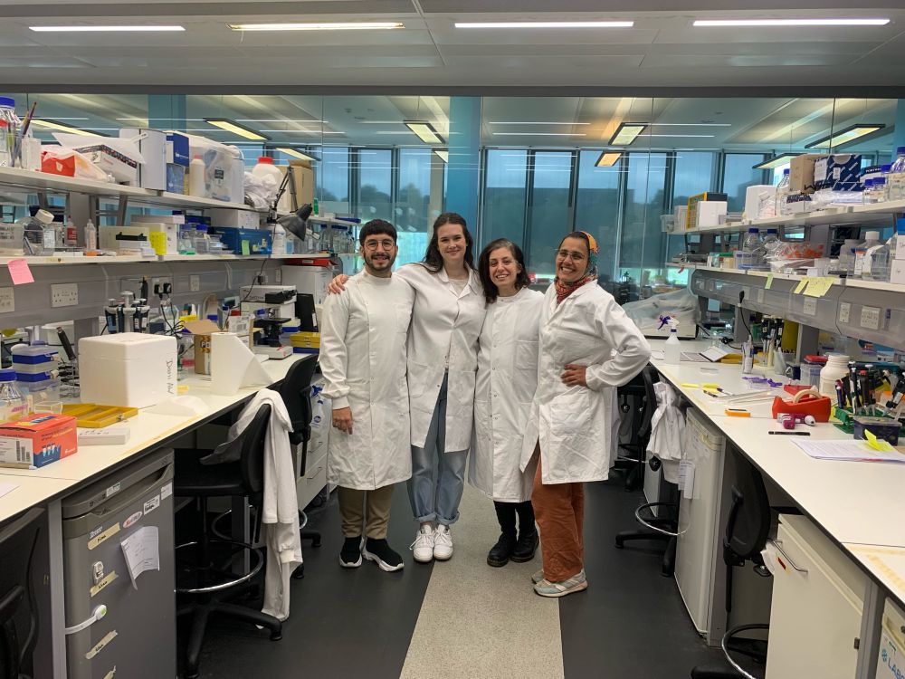 A picture of me with Rebecca Smith, Garyfallia Gouna and Rana Fetit, three of the other postdocs at the Williams lab. We are all wearing our lab coats in the lab, with two long benches full of chemicals and lab tools.