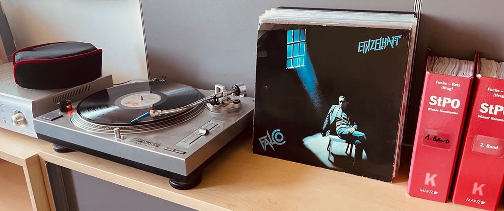Ein Plattenspieler, darauf die Schallplatte "Einzelhaft" des Künstlers Falco deren erstes Lied der Titel "Zu viel Hitze" ist, daneben das Cover der Platte und ganz rechts im Bild zwei Bände des Wiener Kommentars zur Strafprozessordnung.