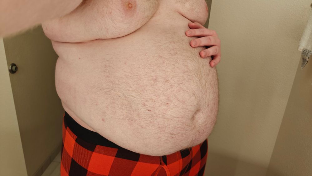 My belly spills over my waistline. This was taken in the hotel I'm currently residing in. I am wearing only red-black checker plaid patterned boxer briefs. One hand rests on the top of my tummy.