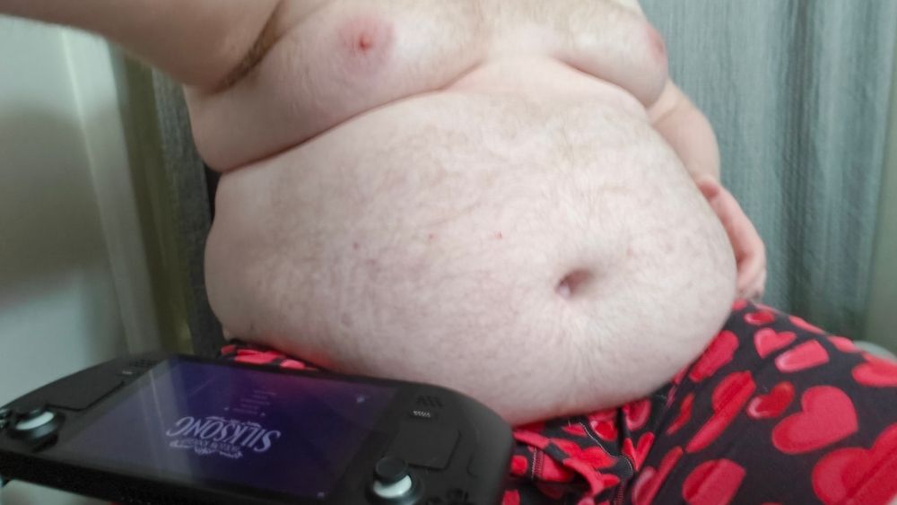 My round belly rests in my lap. I am wearing only heart-patterned boxer briefs. This was taken while dye was setting in my hair. A Steam Deck rests on one leg, and the Hollow Knight Silksong title screen is visible.