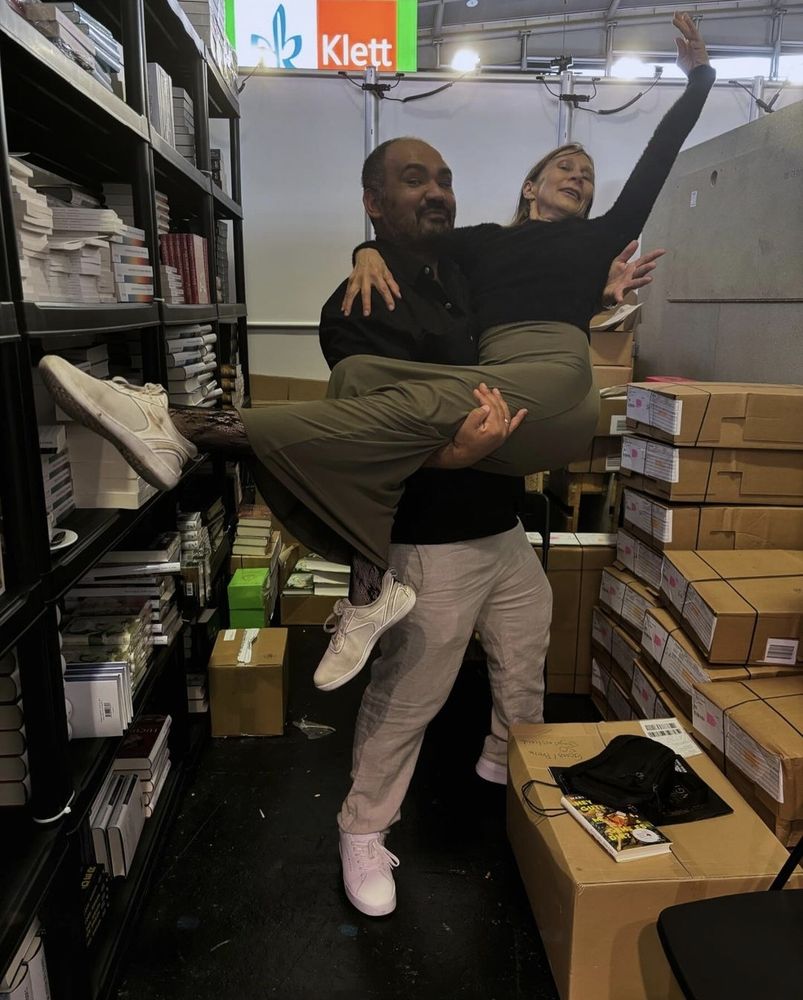 Aufnahme aus dem beengten Lagerraum eines Buchmessestandes. Dinçer Güçyeter trägt Martina Hefter stolz und mit kleinem Augenzwinkern auf Händen, während sie divenhaft theatralisch und ebenso augenzwinkernd einen Arm und ein Bein ausstreckt.