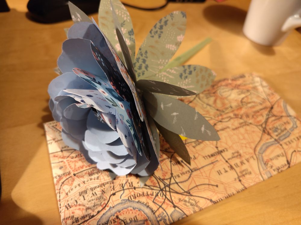 Eine Blume aus mehreren Lagen Origamipapier liegt auf einem selbstgefalteten Umschlag mit Landkartenprint.