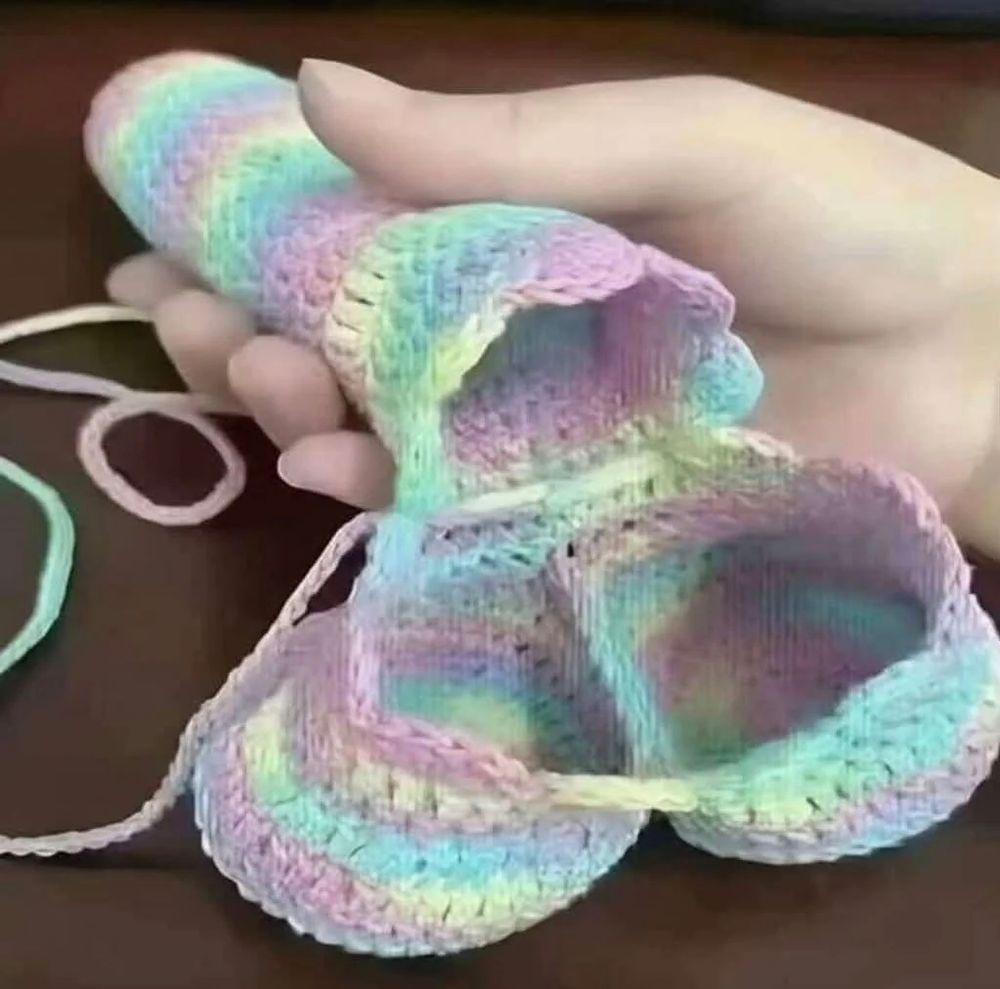 Mão segurando um, ao que tudo indica, agasalho de piroca multicolorido. A peça aparenta ser feita com crochê, possuindo formato fálico e amplo espaço para os testículos. 