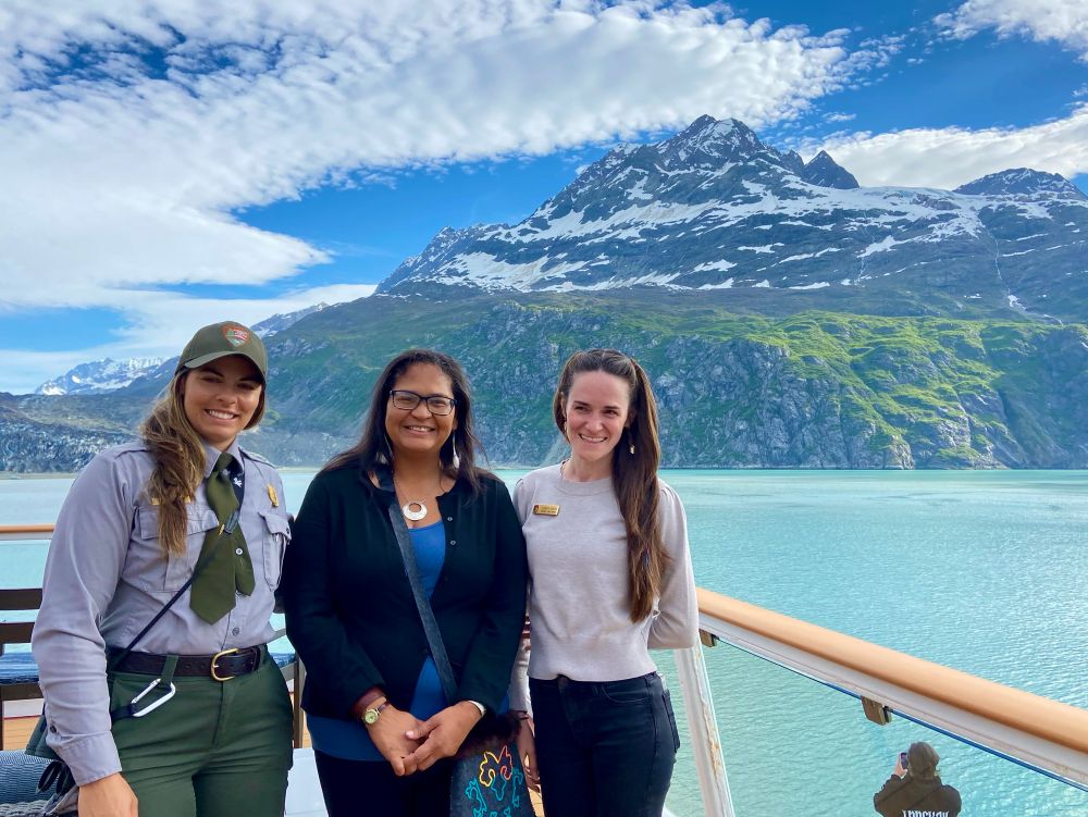 Three women with long sleeves and one in NPS uniform stand in front of a mountain which has some snow and some greenery with blue sky and white clouds and the sea is sort of celadon in color 