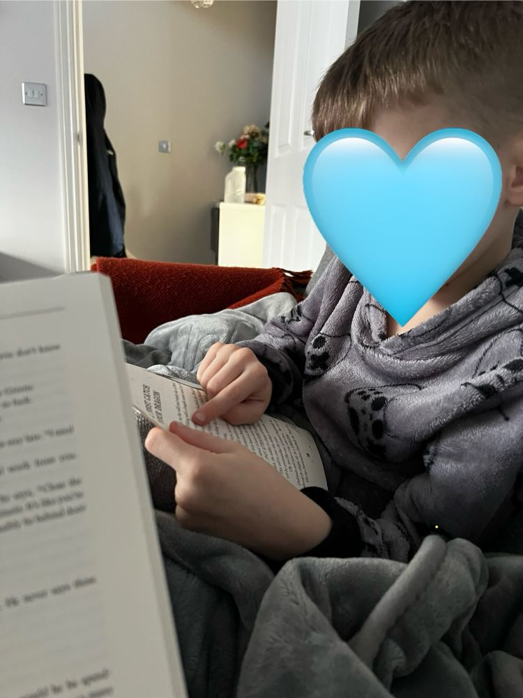 Picture of my son sitting next to me reading a book. We’re sharing a grey blanket. He’s using his finger to track the text. You can see the edge of my book in the image, but the words are not in focus. His face has been hidden by a blue heart.