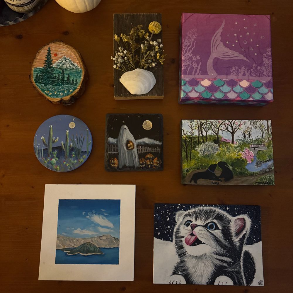 Small art, including a mountain scene painted on a slice of tree, circular painted cacti under a moon, Crater Lake’s island, a black and white cat with its tongue out in the snow, a black dog in woods, a ghost holding a pumpkin, a mermaid tail and dried flowers in a shell. 