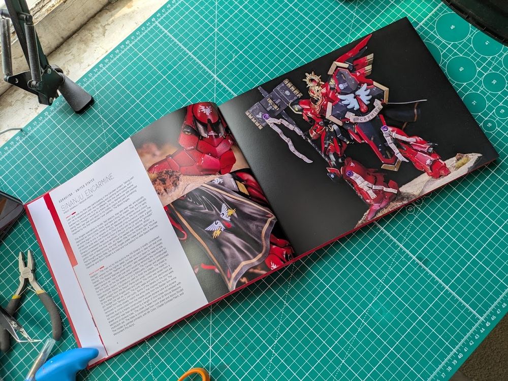 An interior shot of a large hardback book spread open showing 2 pages, on the right is a single large image of a custom gunpla figure painted red. On the left are two smaller images showing details from the right hand page and a column of text. The text is illegible in the image, but it gives the name of the piece and the artists (me) handle on instagram as well as a description of the work done and the theme of the piece.