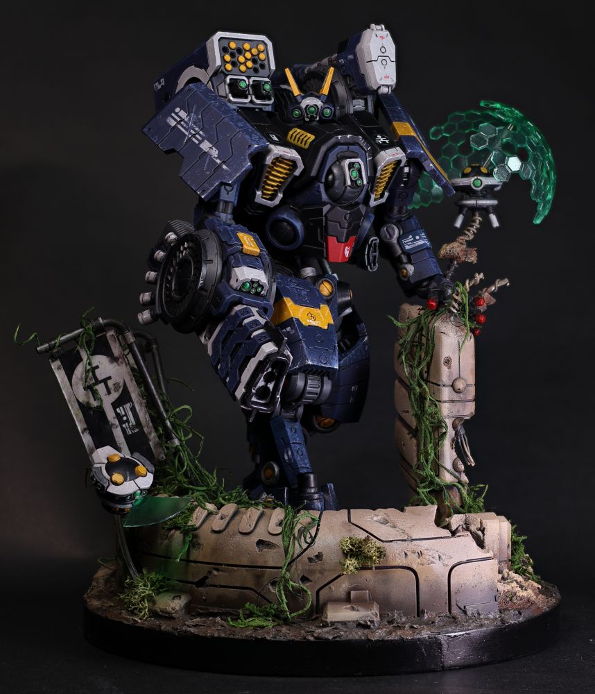 A large blue mech stands amid a crumbling and overgrown ruin. To the left of the image a small drone scans the dilapidated structure. In the upper right a shield drone hovers at the battle suits shoulder.