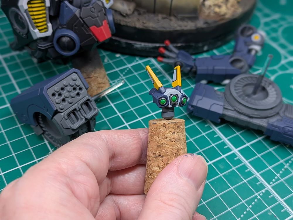 A roboti head mounted to a cork for ease of handling, it is blue with a white central panel and yellow antennae that resemble ears. In the background are more parts of the same kit, currently painted mostly blue and grey